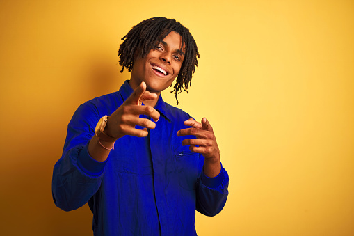 Afro worker man with dreadlocks wearing mechanic uniform over isolated yellow background pointing fingers to camera with happy and funny face. Good energy and vibes.