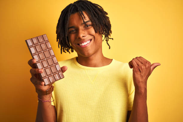 istock Afro american man with dreadlocks eating chocolate bar over isolated yellow background pointing and showing with thumb up to the side with happy face smiling 1175465262