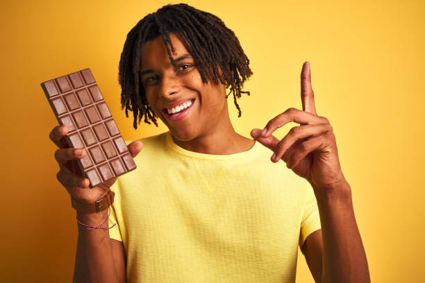 istock Afro american man with dreadlocks eating chocolate bar over isolated yellow background surprised with an idea or question pointing finger with happy face, number one 1175465319