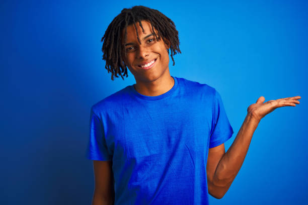istock Afro american man with dreadlocks wearing t-shirt standing over isolated blue background smiling cheerful presenting and pointing with palm of hand looking at the camera. 1175467821