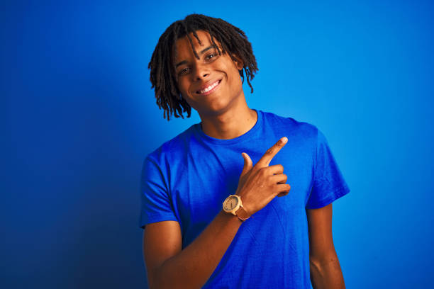 istock Afro american man with dreadlocks wearing t-shirt standing over isolated blue background cheerful with a smile of face pointing with hand and finger up to the side with happy and natural expression on face 1175467832