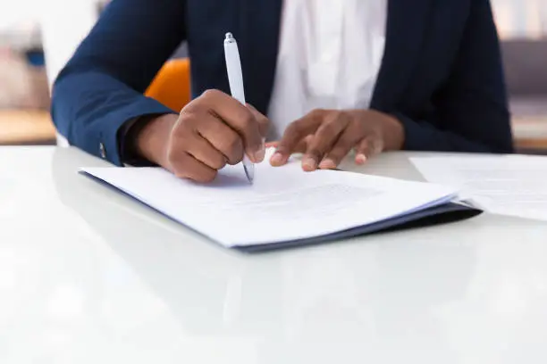 Businesswoman signing contract. African American business woman sitting at table in office, holding pen and writing in document. Legal expertise concept