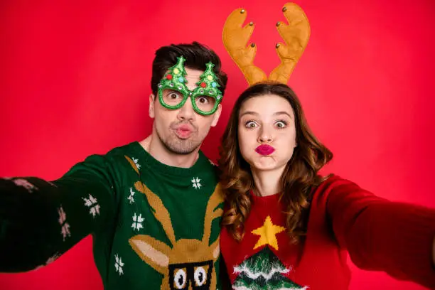 Photo of Photo of carefree lady and guy at newyear costume party making selfies with strange faces wear ugly pullovers isolated red color background