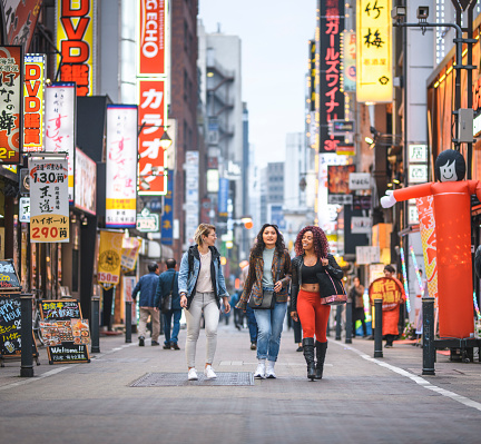 Wide angle view of African, Mongolian, and Caucasian female tourists walking together through Shinjuku in Tokyo at dusk.