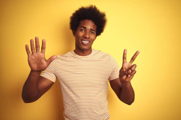 amerikanischer mann mit afro-haar trägt gestreiftes t-shirt steht über isolierten gelben hintergrund zeigt und zeigt mit den fingern nummer sieben, während lächelnd zuversichtlich und glücklich. - beautiful one person men male stock-fotos und bilder