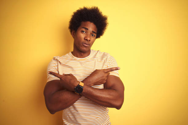 孤立した黄色の背景の上に立つ縞模様のtシャツを着たアメリカ人男性指で両側を指差し、異なる方向は反対 - space to left ストックフォトと画像