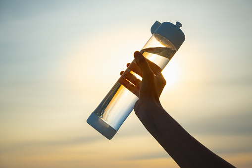 La mano humana sostiene una botella de agua contra el sol poniente. photo