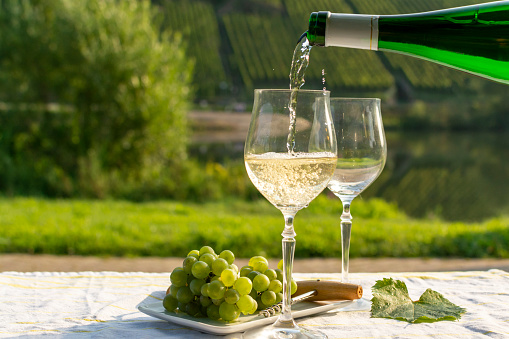 Waiter pouring  German quality white wine riesling, produced in Mosel wine regio from white grapes growing on slopes of hills in Mosel river valley in Germany, bottle and glasses served outside in Mosel valley