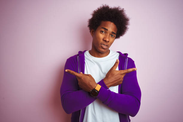 孤立したピンクの背景の上に立つ紫色のスウェットシャツを着た若いアフリカ系アメリカ人男性指で両側を指差し、異なる方向は反対する - space to left ストックフォトと画像