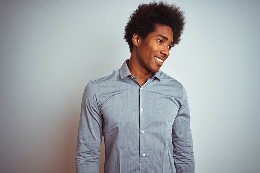 Young african american man with afro hair wearing grey shirt over isolated white background looking away to side with smile on face, natural expression. Laughing confident.