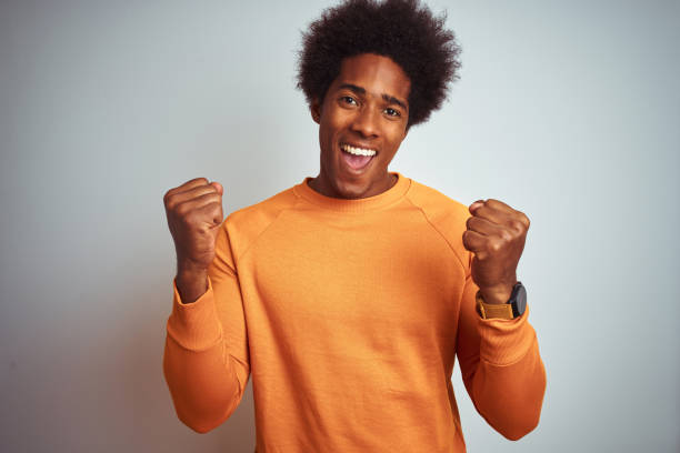 istock Young american man with afro hair wearing orange sweater over isolated white background celebrating surprised and amazed for success with arms raised and open eyes. Winner concept. 1175486830