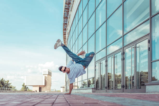 strong sporty man dancing break dance, standing one arm, young guy, free space for text motivation lifestyle, summer city against background glass building window, jeans, sunglasses, a white T-shirt stock photo