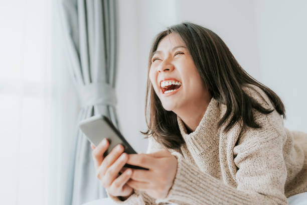Happy woman laughing while lying on the bed with smartphone Happy Asian woman laughing while lying on the bed using smartphone mobile at home Laughing stock pictures, royalty-free photos & images
