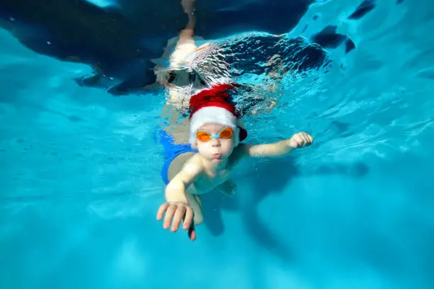 Happy little boy swims underwater in Santa's red hat and swimming glasses. He looks into the camera and blows bubbles. Portrait. Concept. Underwater photography.