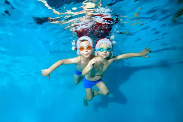 Two little boys swim and pose underwater in red Santa hats and swimming goggles. They hug, look at the camera and smile with their arms outstretched. Christmas celebration. Portrait. Concept.