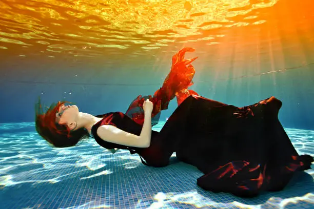 Beautiful young girl lying at the bottom of the pool. She lies with her eyes closed in a burgundy dress, her red hair against the sunlight from the surface. Surrealism. Concept. Landscape orientation.