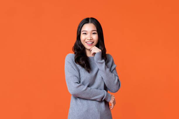 Happy Asian woman smiling with hand on chin Young happy Asian woman smiling with hand on chin isolated on orange studio background natural beauty people photos stock pictures, royalty-free photos & images