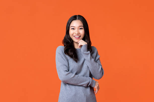 Happy Asian woman smiling with hand on chin Young happy Asian woman smiling with hand on chin isolated on orange studio background asian american model stock pictures, royalty-free photos & images
