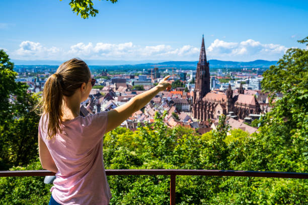 Germany, Young Blonde beauty girl standing above roofs of city freiburg im breisgau historic district and popular muenster cathedral in summertime with blue sky stock photo