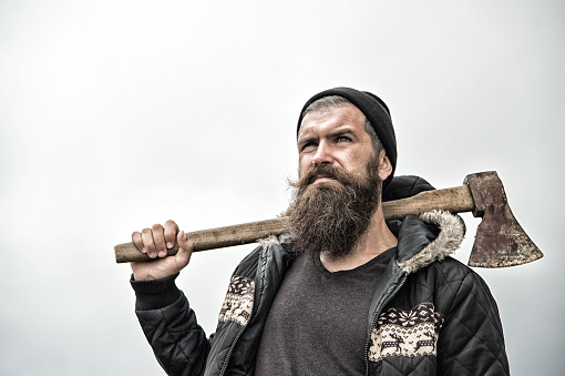 Hipster with beard on serious face carries axe on shoulder sky on background, copy space. Lumberjack brutal and bearded holds axe. Brutal lumberjack concept. Man in hat and warm jacket looks brutally.