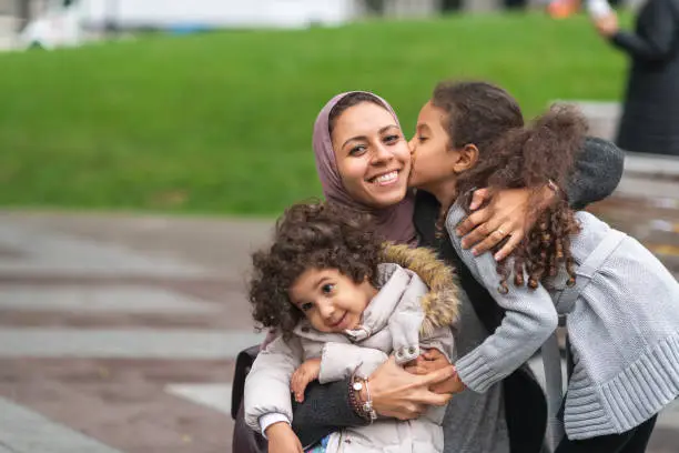 A family of Middle Eastern descent is spending time together. A young mother is playing at the park with her two young daughters. The mother is kneeling down and embracing her children. The happy group is smiling. The older daughter is giving her mom a kiss on the cheek.