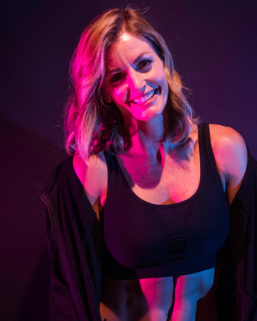 Waist up / one person of 20-29 years old adult beautiful caucasian female / young women standing in front of black background / multi-colored background / colored background wearing sports clothing / sports bra / shorts / running shorts / jacket who is smiling / happy / cheerful / black color / gel effect lighting