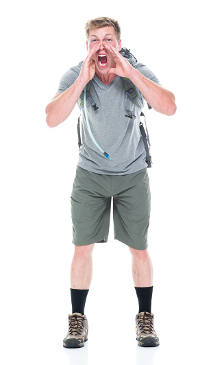 Front view / one person / full length of 20-29 years old caucasian young men / male backpacker hiking / standing in front of white background wearing shorts / backpack / hiking boot / t-shirt who is shouting / screaming / displeased / aggression / angry who is exploration with hands covering mouth and holding bag / explorer