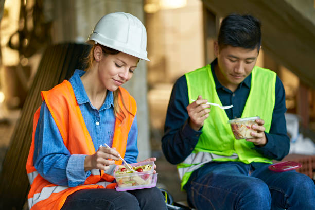 Coworker eating lunch break together at site Multi-ethnic coworker eating lunch break together. Building contractors are sharing lunch while sitting at site. They are wearing reflective clothing. construction lunch break stock pictures, royalty-free photos & images