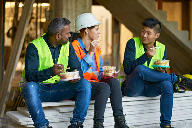 Multi-ethnic coworkers eating lunch at site Multi-ethnic coworkers eating lunch while sitting on planks. Engineers are talking during lunch break. They are wearing protective workwear at construction site. construction lunch break stock pictures, royalty-free photos & images