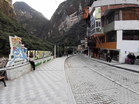 View Of Andes Mountain, Building Exterior, People Taking Picture, Standing, Sitting Down, Walking And Machu Picchu Sign During The Day At Aguas Calientes City Center In Cusco Peru South America
