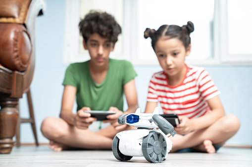 13 years old boy and his 8 years old sister playing with home made robotics at home. They are sitting on floor and controlling robotics with a smartphone. Boy is wearing a green t-shirt while girl is wearing a striped t-shirt. Shot indoor with a full frame mirrorless camera.