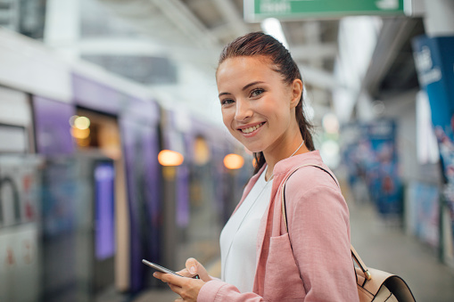 Portrait of beautiful Caucasian young woman standing on metro station and holding a cell phone.