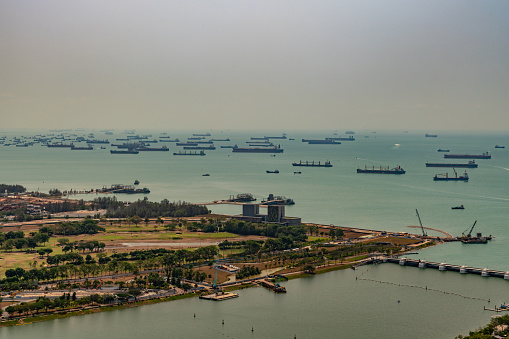 Singapore - March 21, 2019: Birds eye view on tens of ships anchored in Singapore Strait under silver hazy sky. Some coastal land and bay entrance up front.