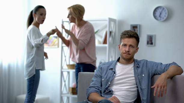 Unhappy man sitting on sofa at home, listening wife quarrelling with mother Unhappy man sitting on sofa at home, listening wife quarrelling with mother mother in law stock pictures, royalty-free photos & images