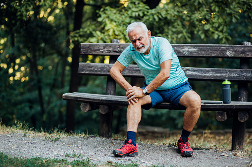 Senior man sitting in the park and holding his painful knee after exercising.