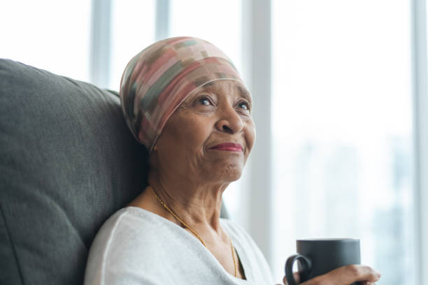 Portrait of a contemplative senior woman with cancer A black senior woman with cancer is wearing a scarf on her head. She is sitting at home in her living room and is holding a cup of tea. She is smiling thoughtfully while looking out a window. She is full of gratitude and hope for recovery. cancer illness stock pictures, royalty-free photos & images