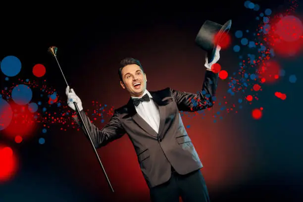 Professional showman wearing suit standing isolated on black and red background holding top hat and pimp cane up smiling excited