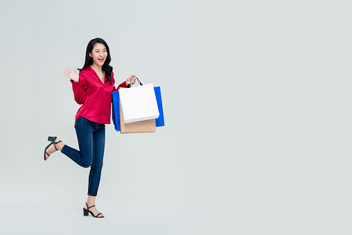 Smiling young Asian girl with shopping bags feeling excited about sale promotion studio shot isolated on light gray background with copy space