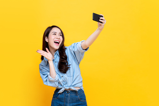 Asian woman wearing casual jean clothes taking selfie and waving hand to smartphone isolated on yellow background.
