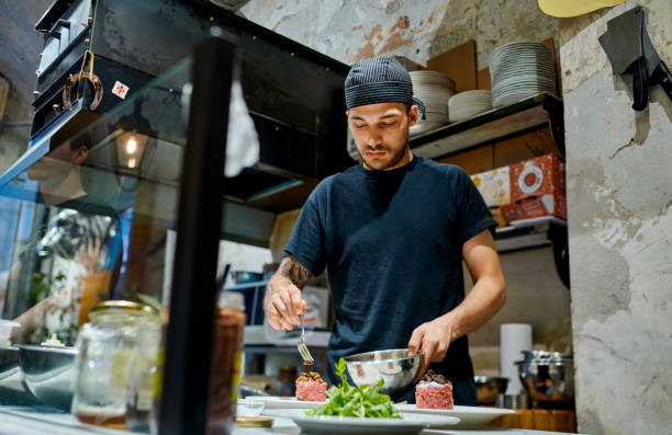 Italian food is so much more than just pasta Shot of a young man preparing a steak tartare dish in a commercial kitchen busy restaurant tables stock pictures, royalty-free photos & images
