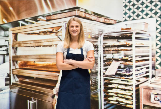 One person / waist up of blond hair caucasian female / young women baker / caterer / retail occupation cooking / preparing food / standing at the bakery / restaurant in the store wearing apron / uniform / t-shirt who is excited / happy / smiling / successful / shouting / confidence and celebration who is working / baking with arms crossed and holding baguette / bread / bun - bread / loaf of bread / small business / food and drink / selling / retail / baked / food una persona / cintura para arriba de pelo rubio mujer caucásica / joven mujeres panadero / catering / venta al por menor de la ocupación de cocina / preparación de alimentos / de pie en la panadería / restaurante en la tienda con delantal / uniforme /