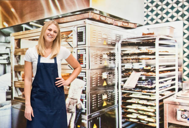 one person / waist up of blond hair caucasian female / young women baker / caterer / retail occupation cooking / preparing food / standing at the bakery / restaurant in the store wearing apron / uniform / t-shirt / shirt and celebration / retail - thank you excitement waist up horizontal imagens e fotografias de stock