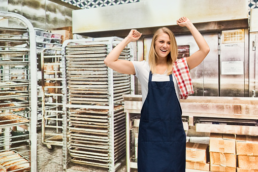 One person / waist up of blond hair caucasian female / young women baker / caterer / retail occupation cooking / preparing food / standing at the bakery / restaurant in the store wearing apron / uniform / t-shirt who is excited / happy / smiling / shouting and celebration and holding or wearing a towel who is working / baking and holding baguette / bread / bun - bread / loaf of bread / small business / food and drink / selling / retail / baked / food