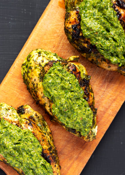 Grilled chimichurri chicken breast on a rustic wooden board on a black surface, top view. Flat lay, overhead, from above. Close-up. Grilled chimichurri chicken breast on a rustic wooden board on a black surface, top view. Flat lay, overhead, from above. Close-up. pesto sauce stock pictures, royalty-free photos & images