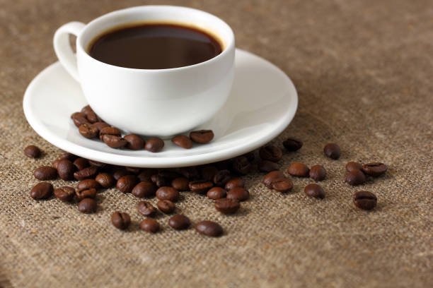 istock White coffee mug on saucer and scattered coffee beans on burlap tablecloth. 1175646602