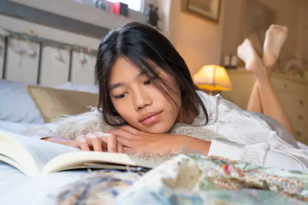 Asian young girl lreading book lying in bed at bedroom