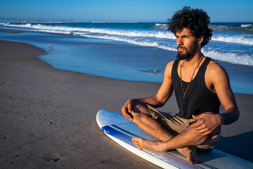 Ethnic young surfer man sitting on surfboard on the beach sand