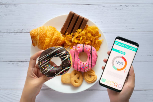 Calories counting and food control concept. woman using Calorie counter application on her smartphone with doughnut in hand and snack , cookies on plate at background Calories counting and food control concept. woman using Calorie counter application on her smartphone with doughnut in hand and snack , cookies on plate at background sugar control stock pictures, royalty-free photos & images