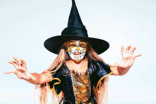 Little girl like a witch in scary yellow-black costume with glitter on white background. Caucasian female model looks scary. Halloween, black friday, sales, autumn holidays concept. The night of fear.