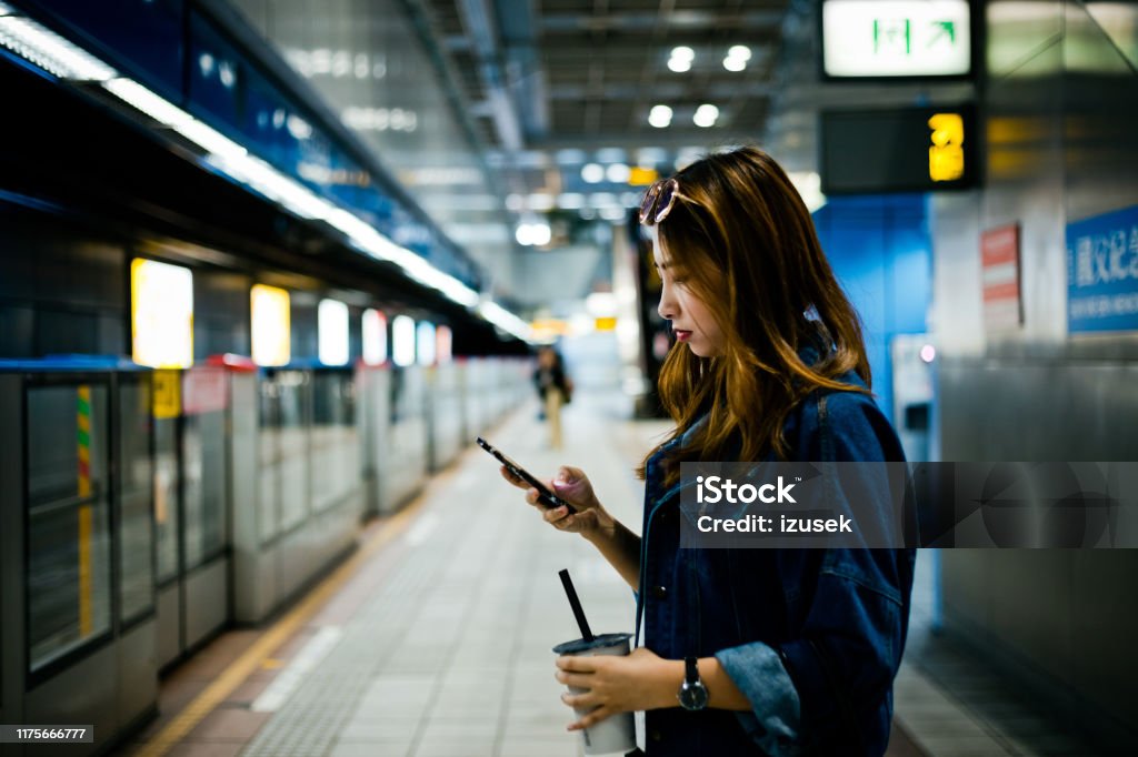Young woman using smart phone at subway station Side view of young woman text messaging with smart phone. Female is standing with disposable cup at subway station. She is wearing denim jacket. 20-24 Years Stock Photo